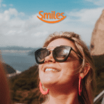 mulher loira com óculos de sol e sorridente com logo Smiles Clube Smiles