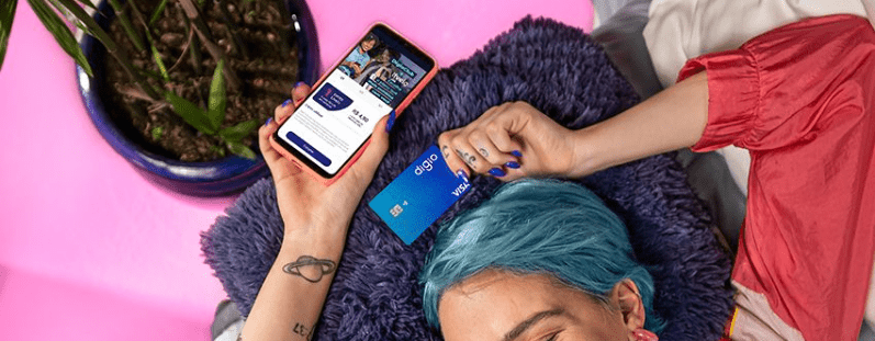 pessoa com cabelo azul deitada em uma cama rosa segurando o cartão Digio e celular  cartão de crédito Digio