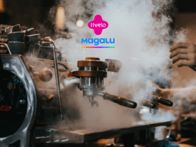 Imagem de uma máquina de café expresso em um ambiente de café, com muita fumaça ao redor, sugerindo a preparação de café fresco. No centro da imagem, sobre a fumaça, estão os logotipos da Livelo (em rosa) e da Magalu (em azul com uma linha colorida abaixo). À direita, uma mão está ajustando a máquina de café. O fundo da imagem é desfocado, destacando a atividade em primeiro plano. 10 pontos livelo