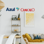 Sala de estar moderna e aconchegante com sofá amarelo e decoração minimalista. Logos da Azul e Camicado destacam uma parceria.