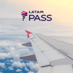 A imagem mostra a asa de um avião com uma ponta vermelha voando acima das nuvens. No canto superior direito, há o logotipo da LATAM Pass. O céu está parcialmente nublado, com o sol iluminando suavemente as nuvens. Pontos Latam Pass
