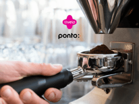 Imagem de uma máquina de café sendo operada, com foco na mão segurando o porta-filtro cheio de café moído. No centro superior da imagem, aparecem os logos da Livelo e do Ponto, indicando uma parceria entre as marcas. Ao fundo, copos desfocados sugerem um ambiente de café ou cozinha. Ganhe até 10 pontos Livelo com o Pontofrio.
