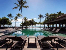 Área da piscina do Kûara Hotel em Arraial D'ajuda, com espreguiçadeiras de frente para a piscina e a praia ao fundo, cercada por palmeiras e vegetação tropical, sob um céu azul claro.
