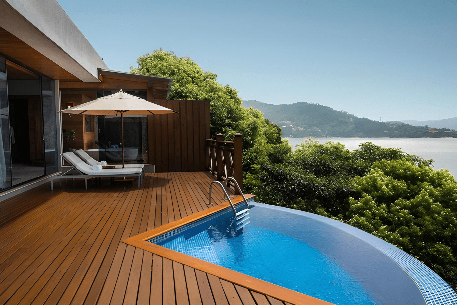 Terraço privativo do Ponta dos Ganchos Exclusive Resort, com piso de madeira e uma piscina de borda infinita que oferece vistas panorâmicas para uma paisagem verdejante e o mar ao fundo. Duas espreguiçadeiras brancas com toalhas enroladas estão dispostas sob um grande guarda-sol, proporcionando sombra e conforto. O ambiente é cercado por vegetação exuberante, criando uma atmosfera de tranquilidade e exclusividade, ideal para relaxamento e apreciação da natureza.