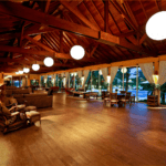 Espaçoso salão de estar no Estaleiro Guest House, com teto alto de madeira e vigas expostas, iluminado por grandes luminárias esféricas. O ambiente é decorado com sofás confortáveis, cadeiras de vime e mesas de madeira, criando uma atmosfera acolhedora e elegante. Grandes janelas de vidro oferecem vistas para a área externa e a piscina, permitindo a entrada de luz natural e proporcionando um ambiente relaxante e sofisticado.