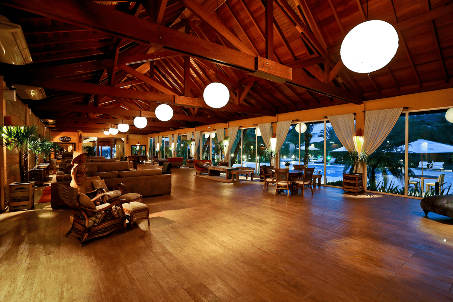 Espaçoso salão de estar no Estaleiro Guest House, com teto alto de madeira e vigas expostas, iluminado por grandes luminárias esféricas. O ambiente é decorado com sofás confortáveis, cadeiras de vime e mesas de madeira, criando uma atmosfera acolhedora e elegante. Grandes janelas de vidro oferecem vistas para a área externa e a piscina, permitindo a entrada de luz natural e proporcionando um ambiente relaxante e sofisticado.