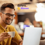 Homem jovem, com pele clara e óculos, usando uma camisa polo amarela, sorri enquanto segura uma xícara de café em uma mão e utiliza um laptop branco. Ele está sentado em uma cafeteria com o fundo desfocado, mostrando luzes e pessoas. No canto superior direito, estão os logotipos das marcas Esfera e Wavee