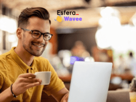 Homem jovem, com pele clara e óculos, usando uma camisa polo amarela, sorri enquanto segura uma xícara de café em uma mão e utiliza um laptop branco. Ele está sentado em uma cafeteria com o fundo desfocado, mostrando luzes e pessoas. No canto superior direito, estão os logotipos das marcas Esfera e Wavee