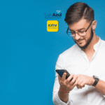 Na imagem, vemos um homem jovem, de barba e óculos, olhando para seu celular com um sorriso no rosto. Ele está vestindo uma camisa branca de mangas compridas e um relógio de pulso. O fundo é de um azul vibrante, destacando o homem em primeiro plano. Acima da cabeça do homem, estão os logotipos do programa de vantagens TudoAzul e do KM de Vantagens, sugerindo uma parceria ou promoção entre as duas marcas. A expressão do homem é de satisfação, provavelmente por estar interagindo com algo positivo em seu celular.