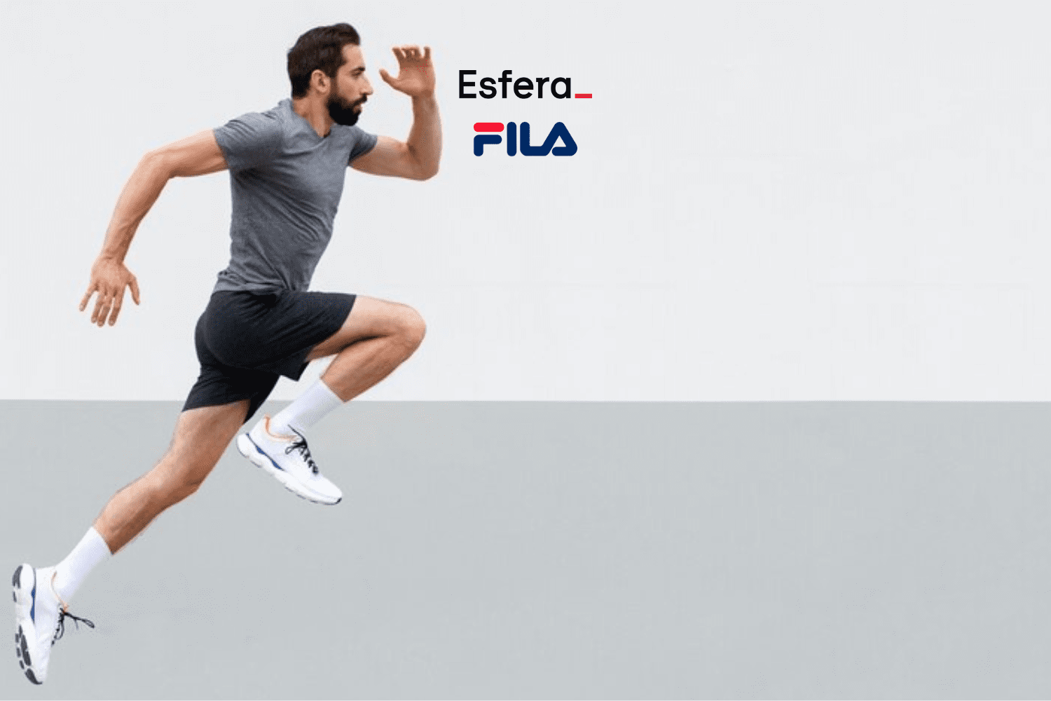 Na imagem, vemos um homem em movimento, capturado enquanto está correndo. Ele está usando uma camiseta cinza escura, shorts pretos e tênis brancos com detalhes coloridos, além de meias brancas. O fundo é dividido em duas partes, sendo a parte superior branca e a inferior cinza claro, criando um contraste que destaca o corredor. Acima da cabeça do homem, estão os logotipos da Esfera e da FILA, sugerindo uma colaboração ou promoção entre as marcas.