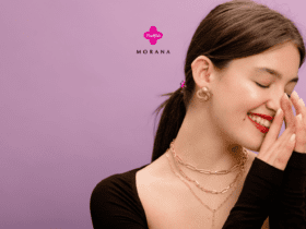 Imagem de uma mulher sorrindo com os olhos fechados, em um fundo lilás. Ela está usando batom vermelho e brincos dourados. A mulher veste uma blusa preta de manga longa e está usando colares dourados. Sua mão esquerda está levantada perto do rosto em um gesto de alegria. No centro da imagem, estão os logos da Livelo e da Morana, sugerindo uma parceria entre as marcas.