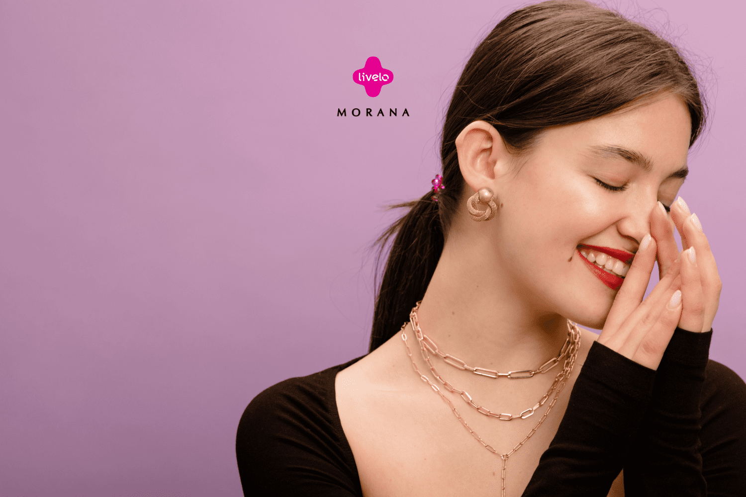Imagem de uma mulher sorrindo com os olhos fechados, em um fundo lilás. Ela está usando batom vermelho e brincos dourados. A mulher veste uma blusa preta de manga longa e está usando colares dourados. Sua mão esquerda está levantada perto do rosto em um gesto de alegria. No centro da imagem, estão os logos da Livelo e da Morana, sugerindo uma parceria entre as marcas.