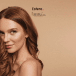 Imagem de uma mulher com cabelo ruivo ondulado em um fundo bege. Ela está sorrindo levemente, olhando para a câmera, e segurando uma mecha do cabelo com a mão direita. Sua pele está iluminada e ela usa maquiagem discreta. No centro superior da imagem, estão os logos da Esfera e da Forever Liss, sugerindo uma parceria entre as marcas.