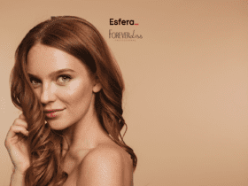Imagem de uma mulher com cabelo ruivo ondulado em um fundo bege. Ela está sorrindo levemente, olhando para a câmera, e segurando uma mecha do cabelo com a mão direita. Sua pele está iluminada e ela usa maquiagem discreta. No centro superior da imagem, estão os logos da Esfera e da Forever Liss, sugerindo uma parceria entre as marcas.