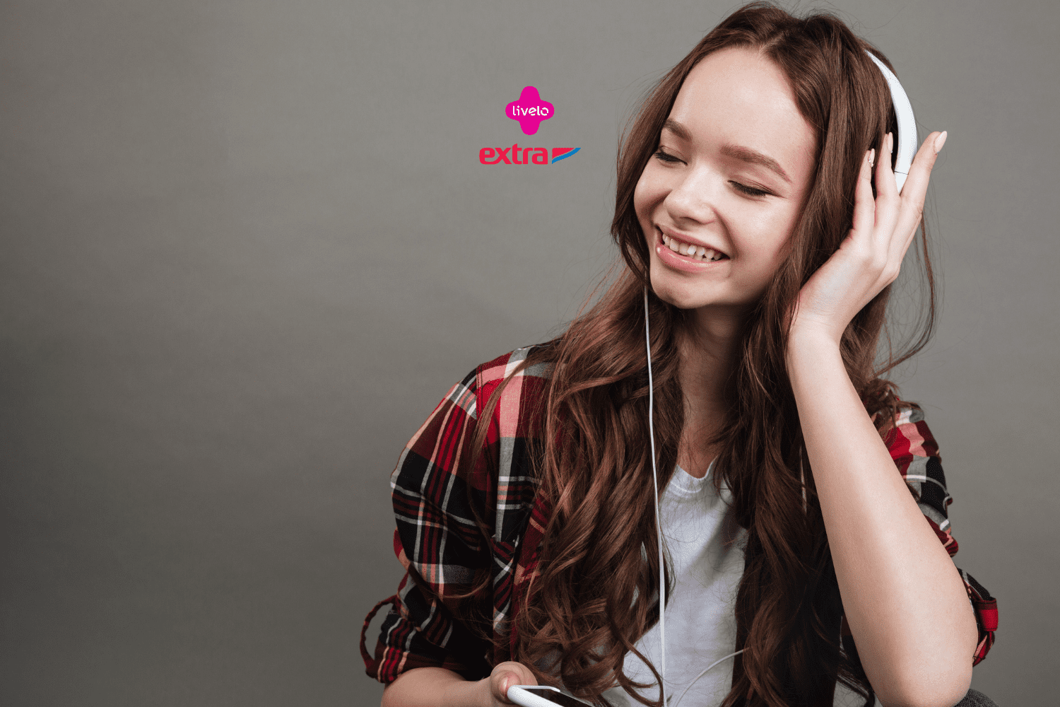 Imagem de uma jovem mulher sorrindo e ouvindo música com fones de ouvido brancos. Ela está usando uma camisa xadrez vermelha e preta por cima de uma camiseta branca. A mulher segura um dispositivo, provavelmente um celular, na mão direita. O fundo é cinza sólido. No centro superior da imagem, estão os logos da Livelo e do Extra, sugerindo uma parceria entre as marcas.