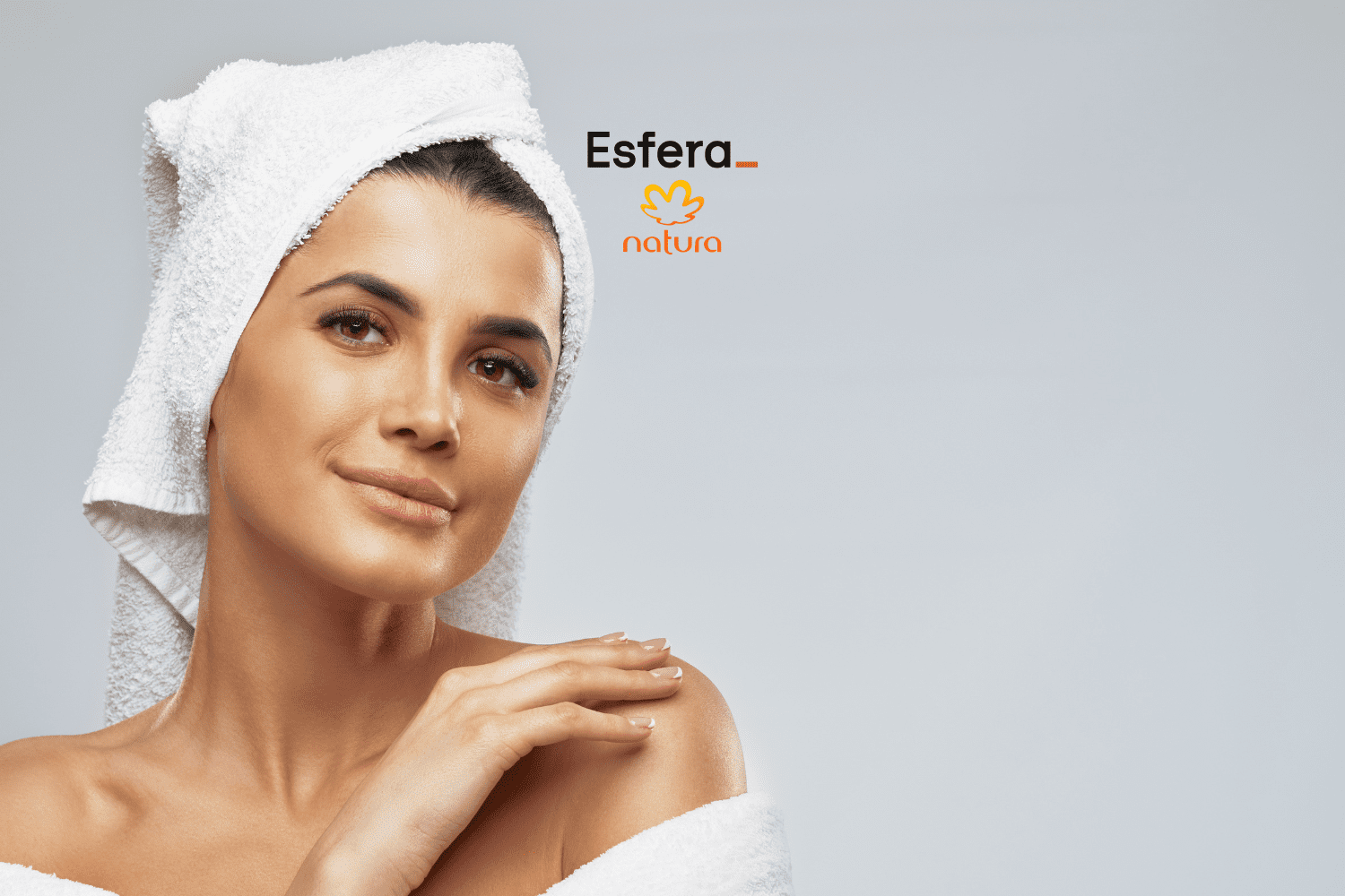 Imagem de uma mulher com uma toalha branca enrolada na cabeça e outra toalha branca envolvendo seus ombros. Ela tem uma expressão serena e está tocando suavemente seu ombro esquerdo com a mão direita. O fundo é cinza claro. No centro superior da imagem, estão os logos da Esfera e da Natura, sugerindo uma parceria entre as marcas.