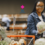 Imagem de uma jovem mulher fazendo compras em um supermercado. Ela está usando óculos, luvas e uma jaqueta jeans. A mulher segura uma cabeça de repolho enquanto olha para ele e sorri. Na frente dela, há um carrinho de compras parcialmente visível com diversos vegetais, incluindo um abacaxi. O fundo mostra prateleiras desfocadas com produtos. No centro superior da imagem, estão os logos da Livelo e do Sam's Club, sugerindo uma parceria entre as marcas