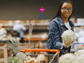 Imagem de uma jovem mulher fazendo compras em um supermercado. Ela está usando óculos, luvas e uma jaqueta jeans. A mulher segura uma cabeça de repolho enquanto olha para ele e sorri. Na frente dela, há um carrinho de compras parcialmente visível com diversos vegetais, incluindo um abacaxi. O fundo mostra prateleiras desfocadas com produtos. No centro superior da imagem, estão os logos da Livelo e do Sam's Club, sugerindo uma parceria entre as marcas