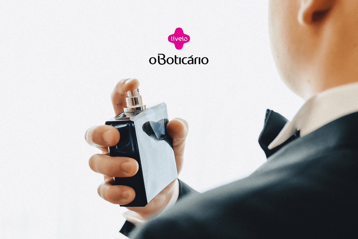 "Imagem de um homem visto de costas, vestindo um terno preto e gravata borboleta. Ele está segurando um frasco de perfume retangular com as mãos, prestes a aplicar o produto. No topo da imagem, há os logos da Livelo e do Boticário.