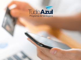 A imagem mostra uma pessoa segurando um smartphone com uma mão e um cartão de crédito com a outra. A imagem está levemente desfocada ao fundo, focando nas mãos e nos objetos que estão sendo segurados. No canto superior direito da imagem, há o logotipo do programa de vantagens "TudoAzul", com um ícone de avião de papel azul.