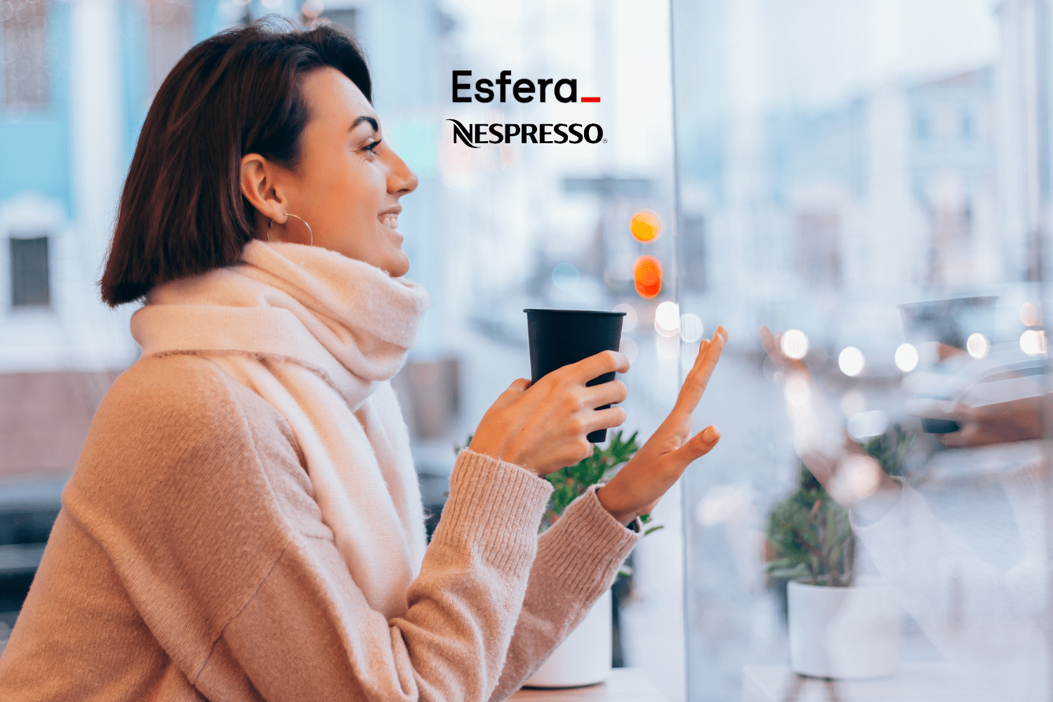Imagem de uma mulher jovem sentada em um café, vista de perfil, usando uma blusa de lã bege e um cachecol branco. Ela segura um copo de café preto com as duas mãos e sorri enquanto olha pela janela. No topo da imagem, há os logos 'Esfera' e 'Nespresso'