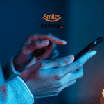 Na imagem, vemos um close-up das mãos de uma pessoa usando um smartphone. A iluminação é predominantemente azul, criando um ambiente moderno e tecnológico. A pessoa parece estar interagindo com o dispositivo, com um dedo pronto para tocar na tela. Ao fundo, há um efeito de desfoque com pontos de luz amarelada. Na parte superior central da imagem, estão os logotipos das empresas Smiles e Esfera.