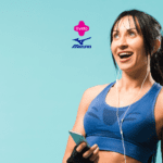 Uma mulher jovem, de cabelos longos e pretos, está sorrindo enquanto segura um celular na mão direita e tem fones de ouvido conectados ao celular. Ela está vestindo um top esportivo azul e luvas de treino pretas sem dedos. O fundo é de cor azul claro. Acima da mulher, estão os logos da "Livelo" e "Mizuno".