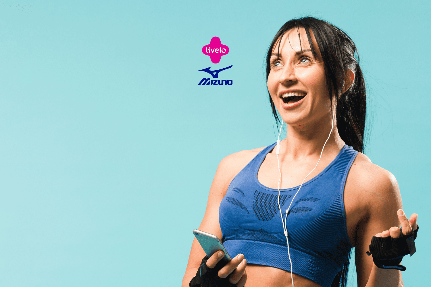 Uma mulher jovem, de cabelos longos e pretos, está sorrindo enquanto segura um celular na mão direita e tem fones de ouvido conectados ao celular. Ela está vestindo um top esportivo azul e luvas de treino pretas sem dedos. O fundo é de cor azul claro. Acima da mulher, estão os logos da "Livelo" e "Mizuno".