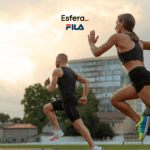 Dois corredores estão em plena corrida ao ar livre. À direita, uma mulher vestindo um top esportivo preto e shorts pretos, com tênis de corrida coloridos, está correndo com determinação. À esquerda, um homem com cabelo curto, vestindo uma regata preta e shorts pretos, também está correndo. O cenário inclui um edifício moderno e árvores ao fundo, com um céu parcialmente nublado ao entardecer. Acima dos corredores, estão os logos da "Esfera" e da "Fila".