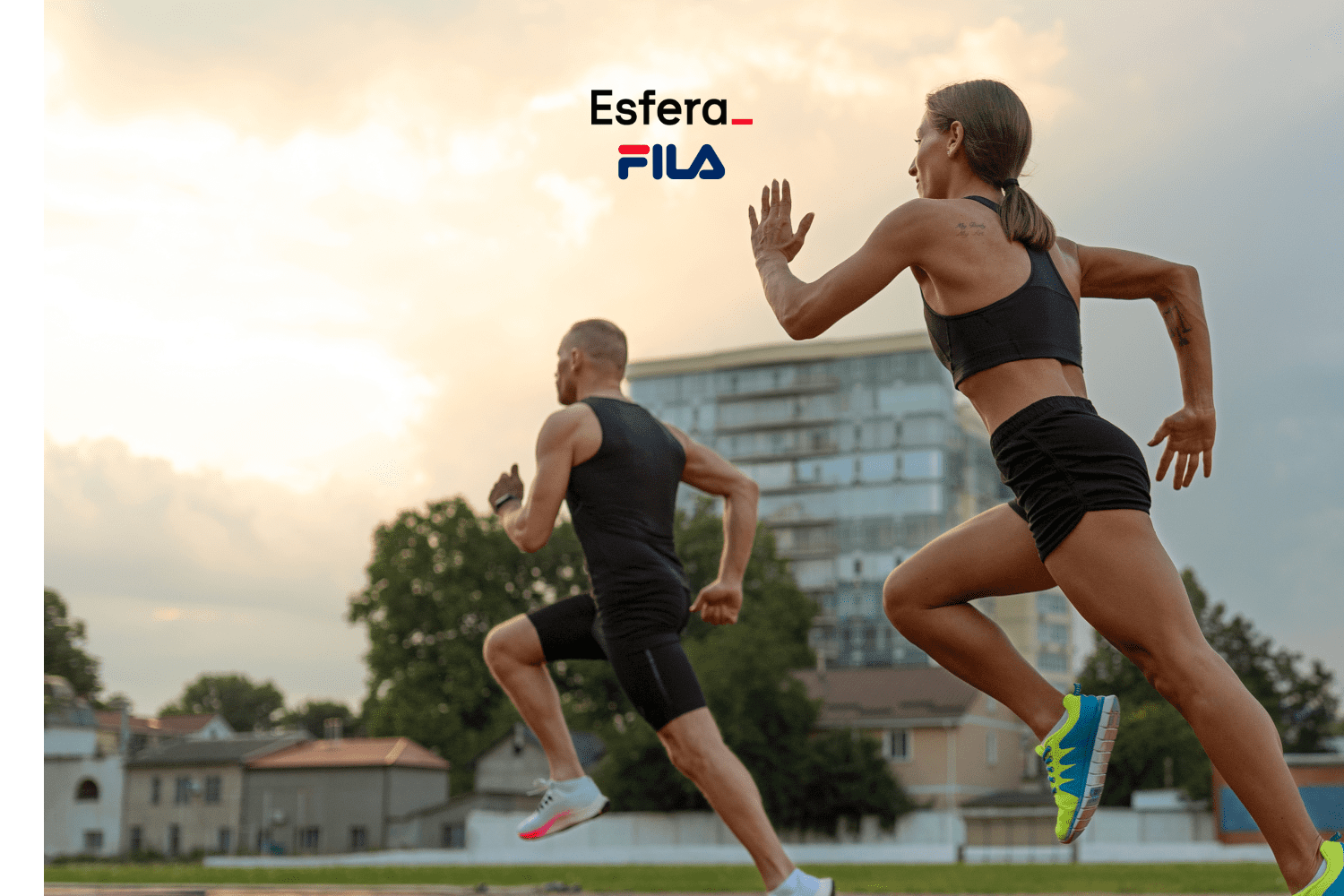 Dois corredores estão em plena corrida ao ar livre. À direita, uma mulher vestindo um top esportivo preto e shorts pretos, com tênis de corrida coloridos, está correndo com determinação. À esquerda, um homem com cabelo curto, vestindo uma regata preta e shorts pretos, também está correndo. O cenário inclui um edifício moderno e árvores ao fundo, com um céu parcialmente nublado ao entardecer. Acima dos corredores, estão os logos da "Esfera" e da "Fila".