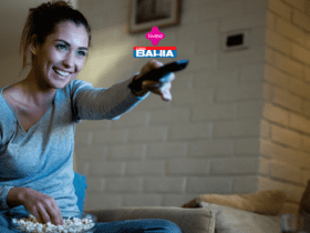 Imagem de uma mulher jovem, sentada em um sofá, assistindo TV. Ela está usando uma blusa de manga comprida cinza e calças confortáveis, enquanto segura um controle remoto em uma mão e uma tigela grande de pipoca na outra. A mulher está sorrindo e parece estar se divertindo. No canto superior direito da imagem, os logotipos da Livelo e das Casas Bahia são visíveis, indicando uma parceria entre as duas marcas.