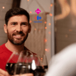 Um homem de barba e cabelo escuro, vestindo um suéter vermelho, está sorrindo enquanto segura uma taça de vinho tinto. Ele está em um ambiente interno, decorado com luzes suaves e festivas. À frente dele, desfocados, estão outras taças de vinho, sugerindo que ele está brindando com outras pessoas. No fundo, há uma decoração aconchegante com luzes decorativas. Acima do homem, estão os logos da "Livelo" e do "Carrefour"