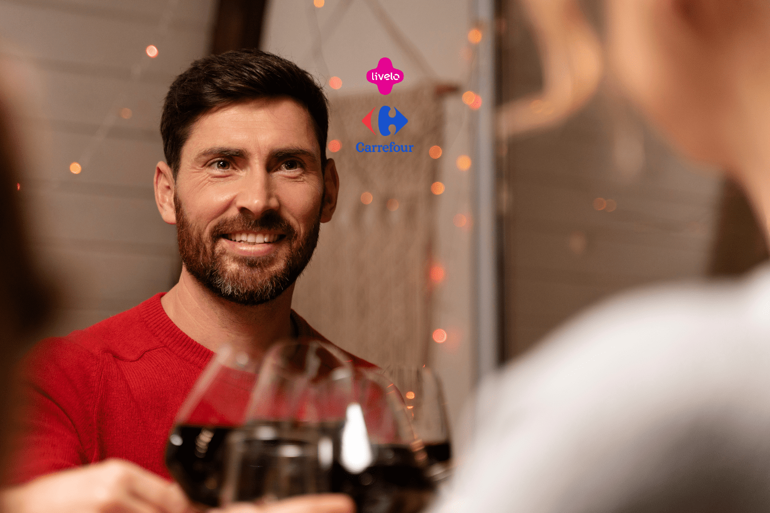 Um homem de barba e cabelo escuro, vestindo um suéter vermelho, está sorrindo enquanto segura uma taça de vinho tinto. Ele está em um ambiente interno, decorado com luzes suaves e festivas. À frente dele, desfocados, estão outras taças de vinho, sugerindo que ele está brindando com outras pessoas. No fundo, há uma decoração aconchegante com luzes decorativas. Acima do homem, estão os logos da "Livelo" e do "Carrefour"