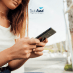 Na foto, uma mulher segura seu celular com ambas as mãos enquanto está sentada ao ar livre, vestindo uma camiseta branca e calça jeans. O logotipo do TudoAzul Programa de Vantagens aparece no canto superior direito da imagem."