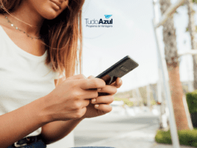 Na foto, uma mulher segura seu celular com ambas as mãos enquanto está sentada ao ar livre, vestindo uma camiseta branca e calça jeans. O logotipo do TudoAzul Programa de Vantagens aparece no canto superior direito da imagem."