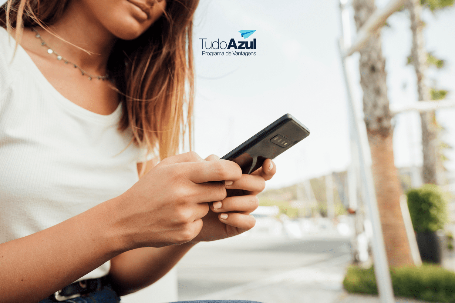 Na foto, uma mulher segura seu celular com ambas as mãos enquanto está sentada ao ar livre, vestindo uma camiseta branca e calça jeans. O logotipo do TudoAzul Programa de Vantagens aparece no canto superior direito da imagem."