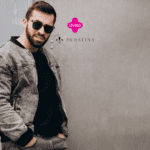 Na imagem, vemos um homem usando óculos escuros e uma jaqueta cinza, posando de forma descontraída em frente a uma parede de cor neutra. Ele veste uma camisa preta por baixo da jaqueta e está com as mãos nos bolsos da calça jeans cinza. O logotipo da Livelo e da Dudalina estão posicionados acima do ombro direito do homem, no canto superior direito da imagem.