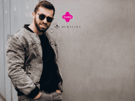 Na imagem, vemos um homem usando óculos escuros e uma jaqueta cinza, posando de forma descontraída em frente a uma parede de cor neutra. Ele veste uma camisa preta por baixo da jaqueta e está com as mãos nos bolsos da calça jeans cinza. O logotipo da Livelo e da Dudalina estão posicionados acima do ombro direito do homem, no canto superior direito da imagem.