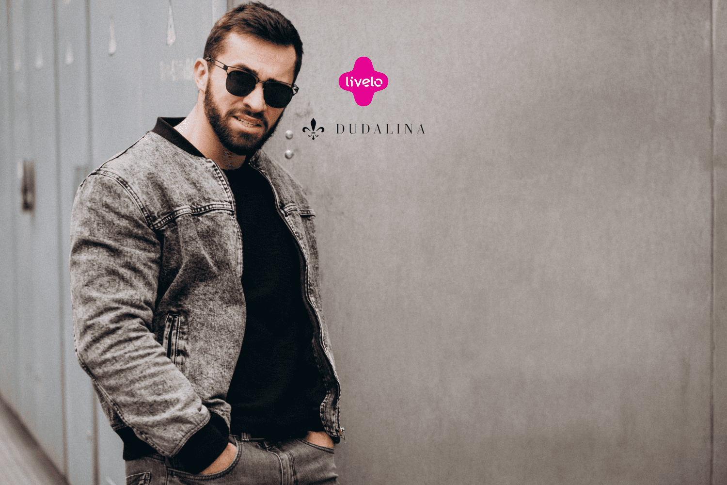 Na imagem, vemos um homem usando óculos escuros e uma jaqueta cinza, posando de forma descontraída em frente a uma parede de cor neutra. Ele veste uma camisa preta por baixo da jaqueta e está com as mãos nos bolsos da calça jeans cinza. O logotipo da Livelo e da Dudalina estão posicionados acima do ombro direito do homem, no canto superior direito da imagem.