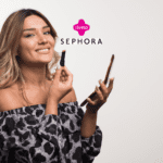 Na foto, uma mulher sorridente aplica batom enquanto segura um celular na outra mão. Ela veste uma blusa ombro a ombro com estampa de leopardo em tons de preto e cinza. Os logotipos da Livelo e Sephora aparecem no topo da imagem."