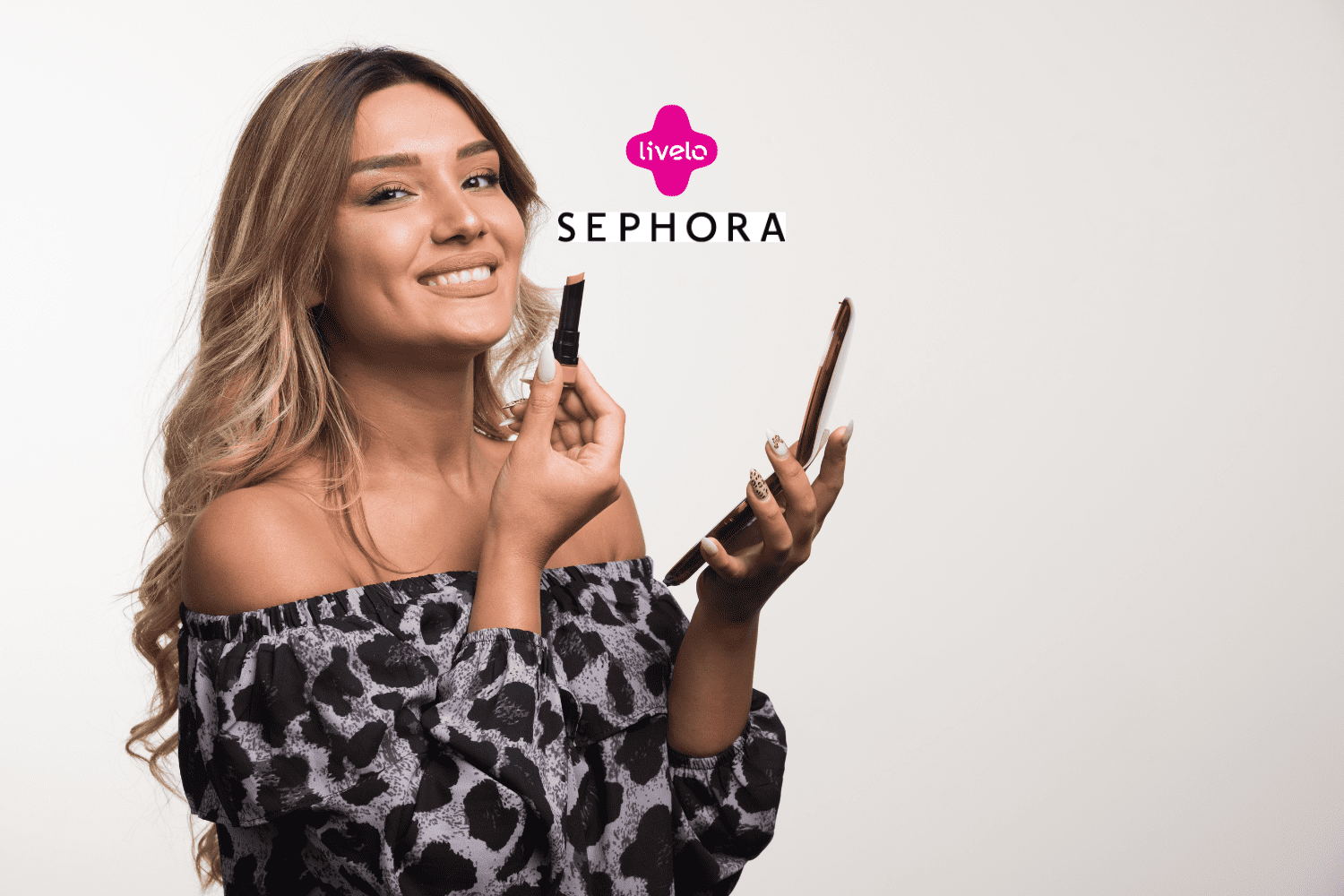 Na foto, uma mulher sorridente aplica batom enquanto segura um celular na outra mão. Ela veste uma blusa ombro a ombro com estampa de leopardo em tons de preto e cinza. Os logotipos da Livelo e Sephora aparecem no topo da imagem."