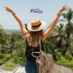 Na foto, uma mulher de costas, com os braços levantados, usa um chapéu de palha e uma blusa preta, enquanto aprecia uma paisagem cheia de palmeiras e vegetação exuberante. Ao fundo, o céu azul claro e o logotipo da LATAM Pass no topo da imagem.
