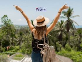 Na foto, uma mulher de costas, com os braços levantados, usa um chapéu de palha e uma blusa preta, enquanto aprecia uma paisagem cheia de palmeiras e vegetação exuberante. Ao fundo, o céu azul claro e o logotipo da LATAM Pass no topo da imagem.