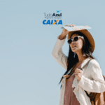 Uma mulher jovem está em um ambiente ao ar livre sob um céu claro e azul. Ela está vestindo um chapéu de palha, óculos de sol grandes e brancos, uma blusa leve de cor creme e um vestido rosa claro. A mulher segura um mapa acima de sua cabeça com a mão direita, como se estivesse protegendo-se do sol, e carrega uma mochila marrom nas costas. No topo da imagem, estão os logotipos "TudoAzul Programa de Vantagens" e "CAIXA" em cores azul e laranja,