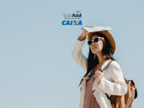 Uma mulher jovem está em um ambiente ao ar livre sob um céu claro e azul. Ela está vestindo um chapéu de palha, óculos de sol grandes e brancos, uma blusa leve de cor creme e um vestido rosa claro. A mulher segura um mapa acima de sua cabeça com a mão direita, como se estivesse protegendo-se do sol, e carrega uma mochila marrom nas costas. No topo da imagem, estão os logotipos "TudoAzul Programa de Vantagens" e "CAIXA" em cores azul e laranja,