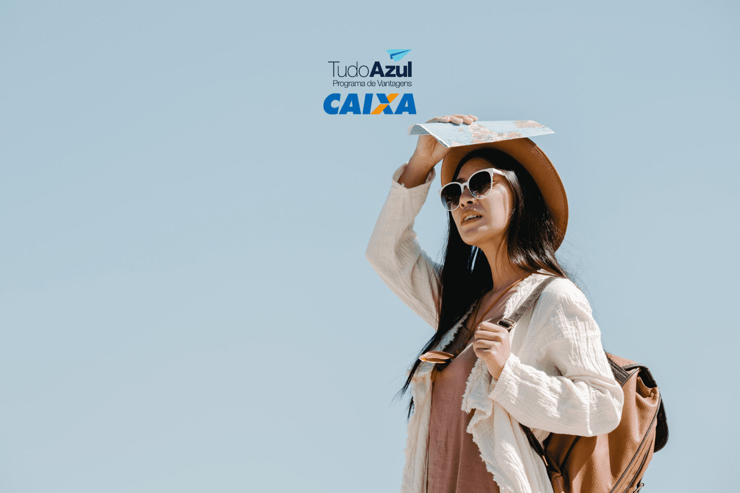 Uma mulher jovem está em um ambiente ao ar livre sob um céu claro e azul. Ela está vestindo um chapéu de palha, óculos de sol grandes e brancos, uma blusa leve de cor creme e um vestido rosa claro. A mulher segura um mapa acima de sua cabeça com a mão direita, como se estivesse protegendo-se do sol, e carrega uma mochila marrom nas costas. No topo da imagem, estão os logotipos "TudoAzul Programa de Vantagens" e "CAIXA" em cores azul e laranja,