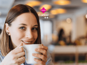 Uma mulher jovem com cabelos castanhos claros e lisos, que vão até os ombros, está sentada em um ambiente de café. Ela sorri levemente enquanto segura uma xícara branca de café próxima ao rosto, parecendo saborear a bebida. A mulher está usando brincos pequenos e uma camisa azul clara. O fundo da imagem está desfocado, mostrando luzes suaves e o interior do café com um ambiente acolhedor. No topo da imagem, estão os logotipos "livelo" em rosa e "Coffee ++