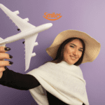 "Uma mulher jovem, com cabelos castanhos e longos, está sorrindo e segurando um avião de brinquedo branco. Ela veste um chapéu de palha e um suéter branco sobre uma blusa preta, posando contra um fundo roxo. Acima de sua cabeça, há o logotipo da Smiles, uma empresa de programa de milhas.