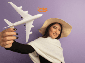 "Uma mulher jovem, com cabelos castanhos e longos, está sorrindo e segurando um avião de brinquedo branco. Ela veste um chapéu de palha e um suéter branco sobre uma blusa preta, posando contra um fundo roxo. Acima de sua cabeça, há o logotipo da Smiles, uma empresa de programa de milhas.