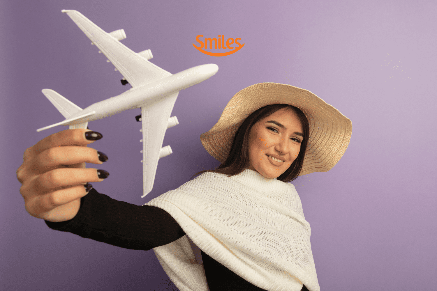"Uma mulher jovem, com cabelos castanhos e longos, está sorrindo e segurando um avião de brinquedo branco. Ela veste um chapéu de palha e um suéter branco sobre uma blusa preta, posando contra um fundo roxo. Acima de sua cabeça, há o logotipo da Smiles, uma empresa de programa de milhas.